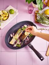 Person seasoning salmon in a pan on a pink tiled table with lemons and herbs.